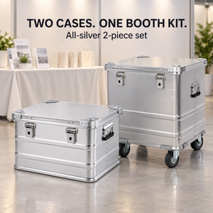 All-silver two-piece aluminum trade show storage set: medium case without wheels and taller case with four-inch casters beside an exhibition booth